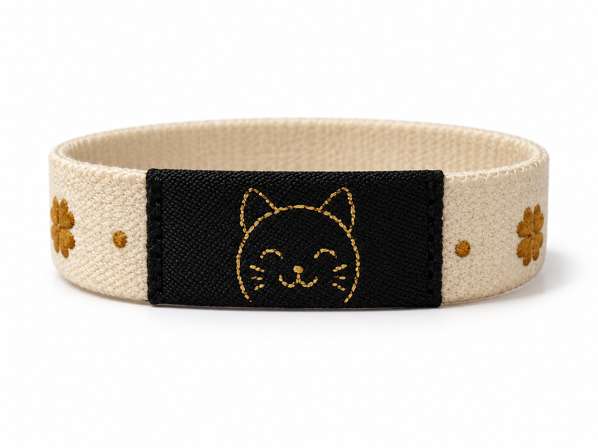 A fabric wristband with a beige woven strap, a central black panel featuring a gold embroidered cat face, and small gold floral details on either side, shown on a white background.