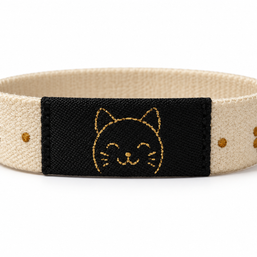 A fabric wristband with a beige woven strap, a central black panel featuring a gold embroidered cat face, and small gold floral details on either side, shown on a white background.